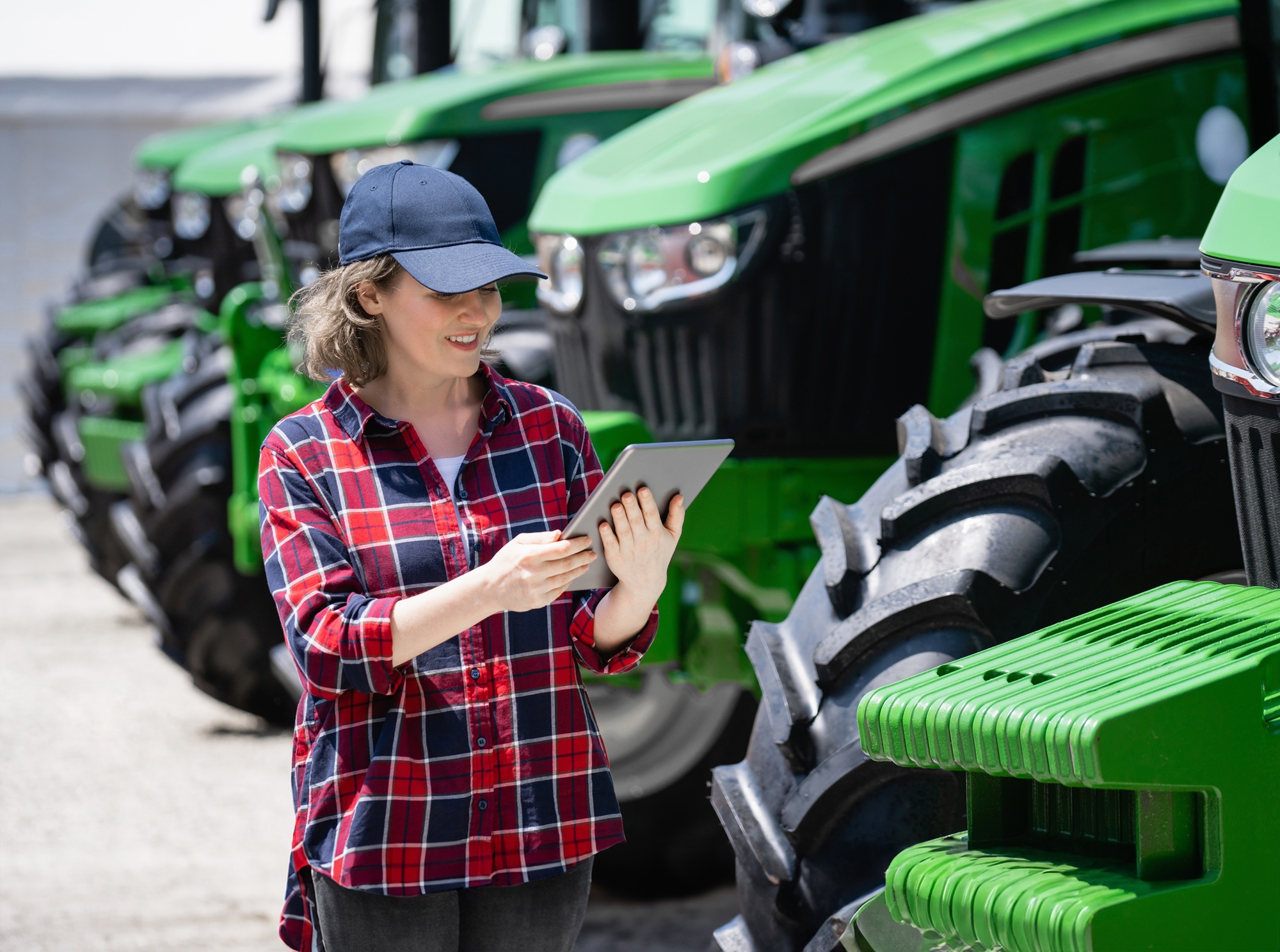 Rolniczka z tabletem cyfrowym na tle ciągników rolniczych.