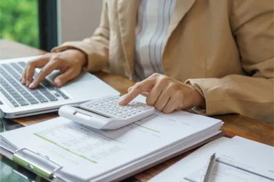 Pracownik liczy na kalkulatorze, pod spodem ma stos dokumentów, jedną ręką korzysta z laptopa
